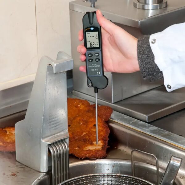 A person wearing a white jacket is using a digital food thermometer to check the temperature of breaded food in a commercial kitchen. The food is placed on a metal surface near a deep fryer. The thermometer displays a temperature reading of 82.5°C.