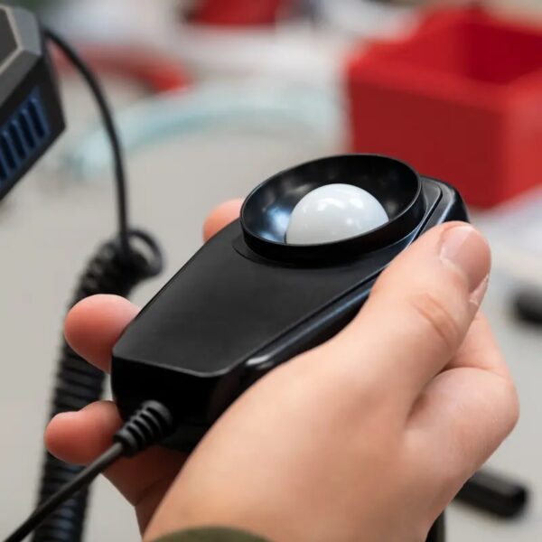 A person holds a black handheld light meter with a circular white sensor. The device has a cord connected to it, and the background shows an out-of-focus work environment with various objects.