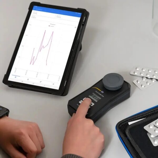 A person uses a handheld medical device with a digital display, next to a tablet showing a line graph. The person is pressing a button on the device, which is connected to the tablet. Nearby, there are packaged pills and a carrying case.