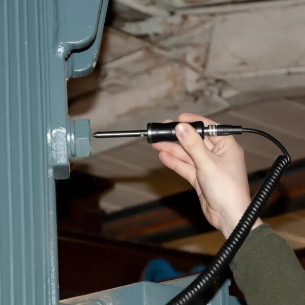 A person's hand is holding a black and silver handheld device with a coiled wire, pointing it towards a large metal bolt on a piece of machinery. The background is an industrial environment with metal beams and equipment.