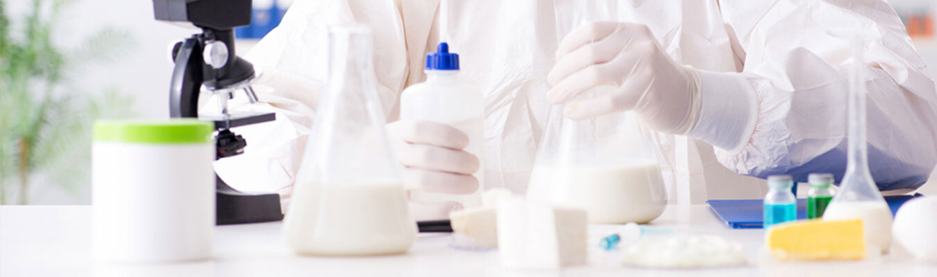 A scientist wearing protective gear works in a laboratory. They are holding a flask and a dropper bottle while other glassware, chemical substances, and a microscope are visible on the table. The scene suggests a focus on scientific research and experiments.