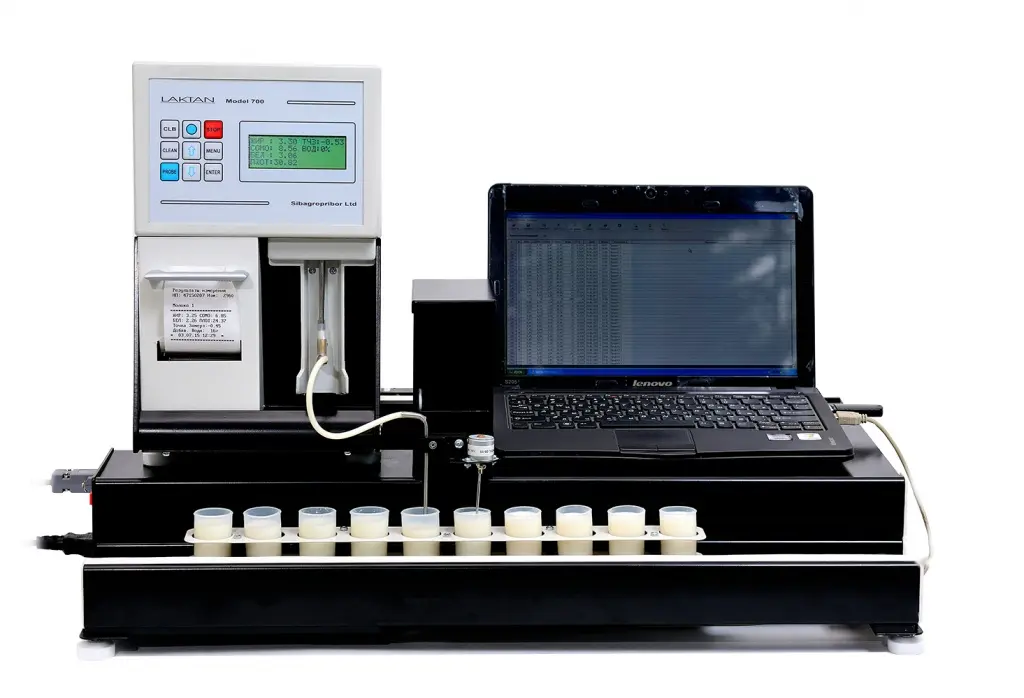 A laboratory setup featuring an LACTAN Mast T10B device connected to a Lenovo laptop. The device has a display screen and attached printer, with several small sample containers in the forefront. The laptop screen shows data in a spreadsheet format.