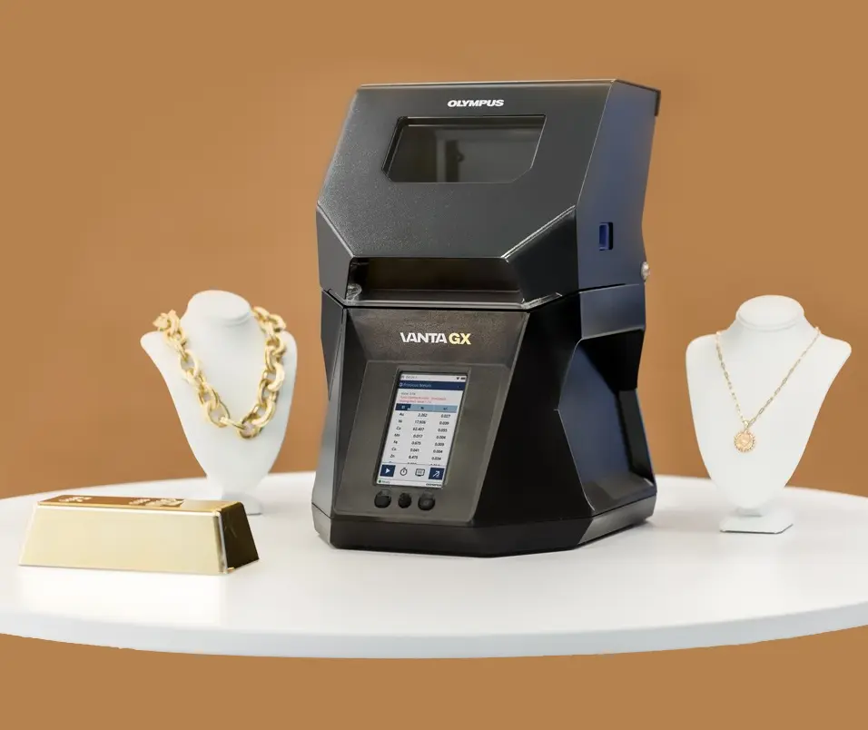 A black Olympus Vanta GX device sits on a white circular table surrounded by various jewelry items: a gold bar, a gold chain necklace, and a pendant necklace on a white bust display. The background is a solid brown color.