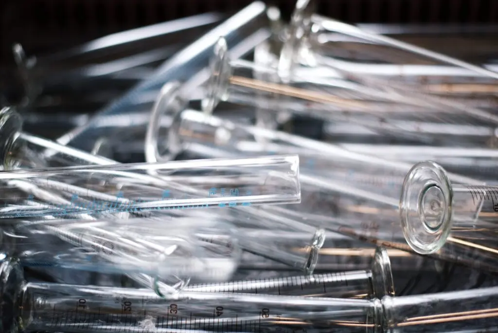 A close-up of several empty glass graduated cylinders lying haphazardly in a pile. The cylinders are transparent, with measurement markings visible on their surfaces. The scene suggests a laboratory environment.