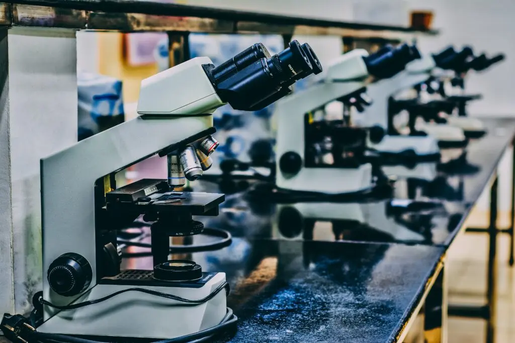 Four microscopes are lined up on a black laboratory countertop. The microscopes have a white body with multiple lenses and black eyepieces. The background is slightly blurred, suggesting a lab setting.