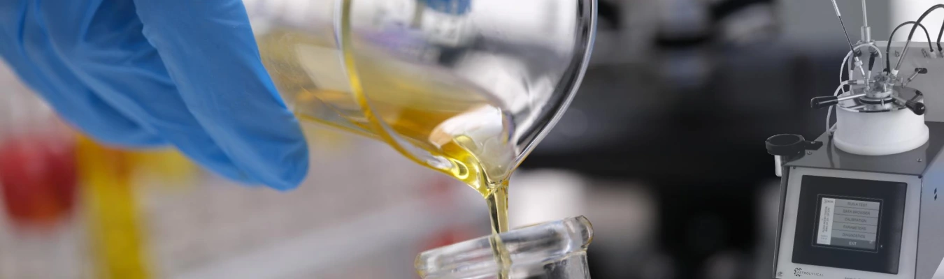 A gloved hand pours yellow liquid from a laboratory flask into another container. On the right, there is a scientific instrument with digital controls and a display screen. The background is blurred lab equipment.